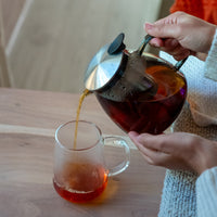 A person pours hot loose leaf tea from the Stormy Glass Teapot with a stainless steel infuser into a clear glass mug on a wooden table, with a cozy blanket partially in view.