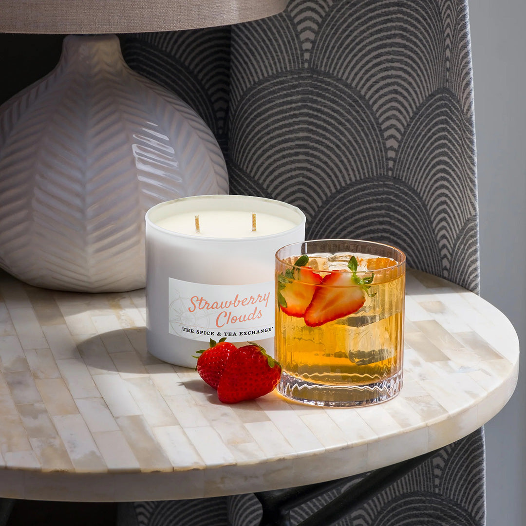 Candle labeled 'Strawberry Clouds' next to a glass of iced tea with strawberries on a marble surface.