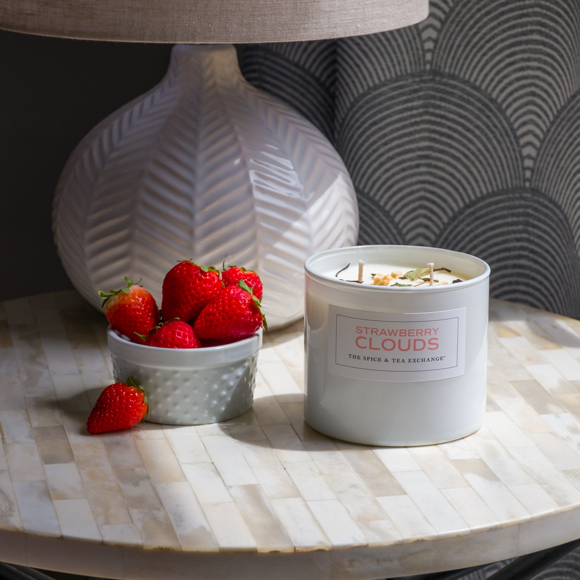A white lamp, a small white bowl of strawberries, and a Strawberry Clouds Candle sit on a round, tiled table next to a patterned wall.