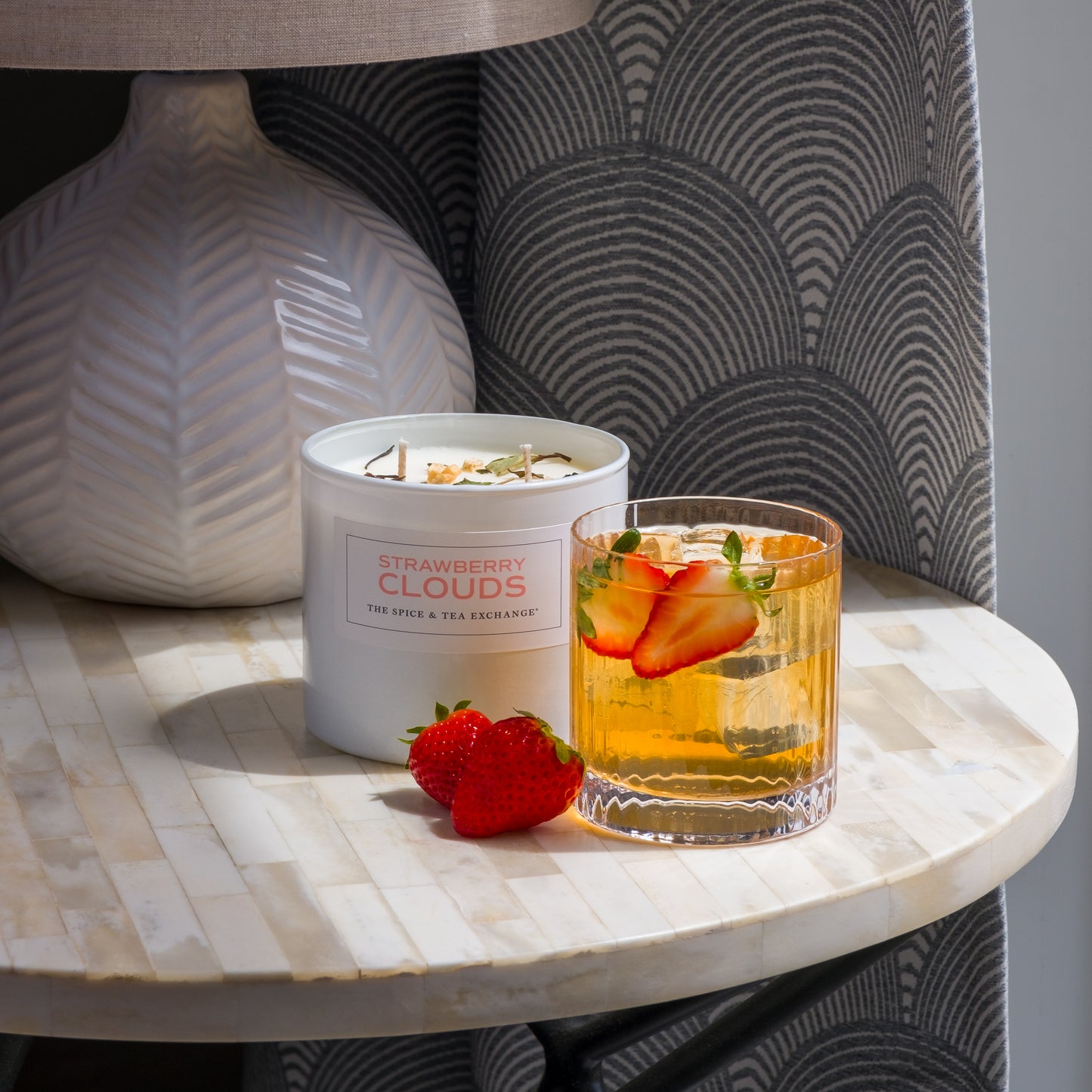 A round marble table with a lamp, a hand-crafted Strawberry Clouds Candle, a glass of iced drink garnished with strawberry slices and herbs, and two fresh strawberries.