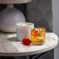 A round marble table with a lamp, a hand-crafted Strawberry Clouds Candle, a glass of iced drink garnished with strawberry slices and herbs, and two fresh strawberries.