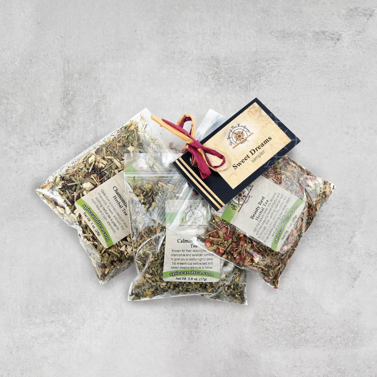 Three clear plastic bags of assorted teas from the Sweet Dreams Sampler are arranged on a grey surface, with a labeled card and ribbon attached.