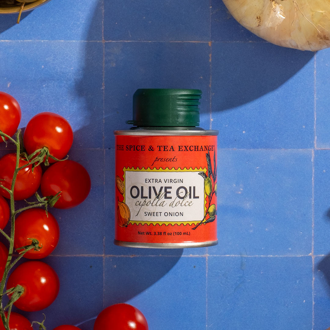 Red can of Sweet Onion Extra Virgin Olive Oil - Mini l with a green lid on a blue tiled surface with tomatoes and an onion.