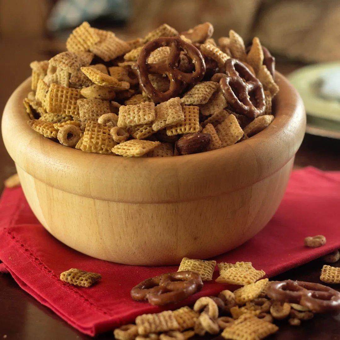 A wooden bowl filled with a snack mix of cereal squares, pretzels, and nuts sits on a red napkin, with a few pieces scattered on the table nearby.