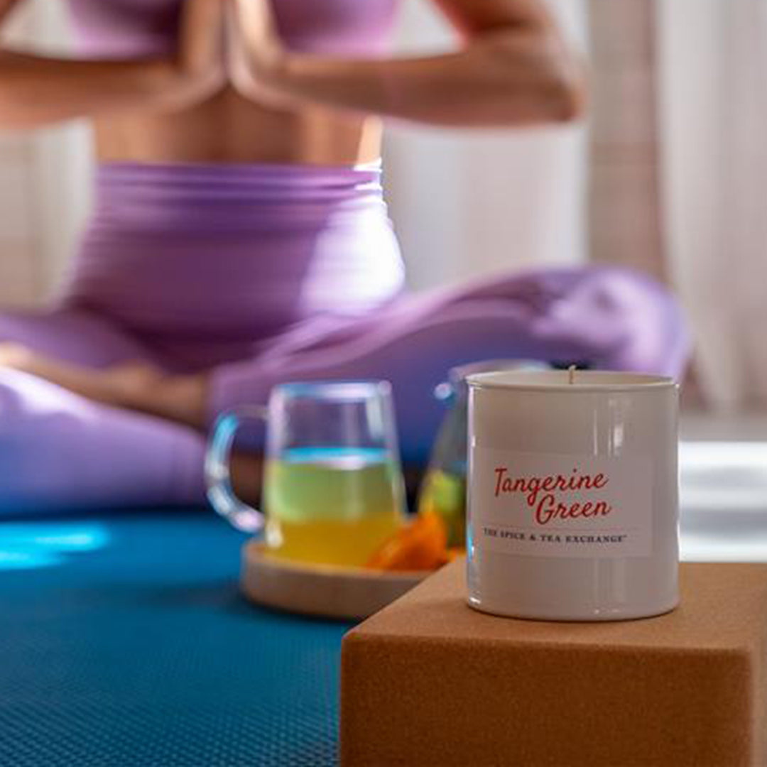 Person in purple outfit meditating with a 'Tangerine Green' candle and mug on a table.