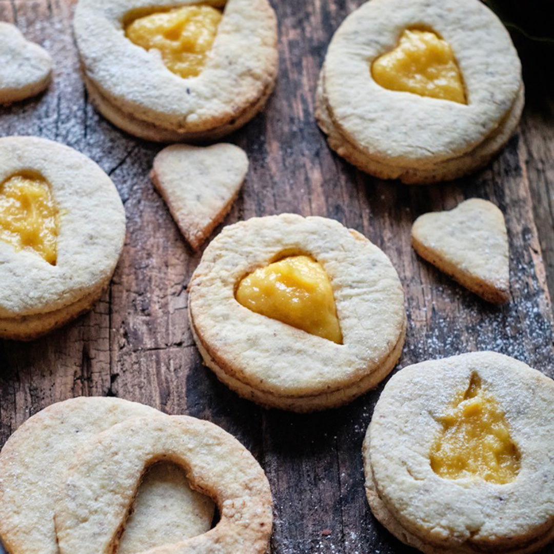 Round sandwich cookies with heart-shaped cutouts are filled with Tangerine Whipped Honey and arranged on a rustic wooden surface, with scattered heart-shaped cookie pieces nearby.