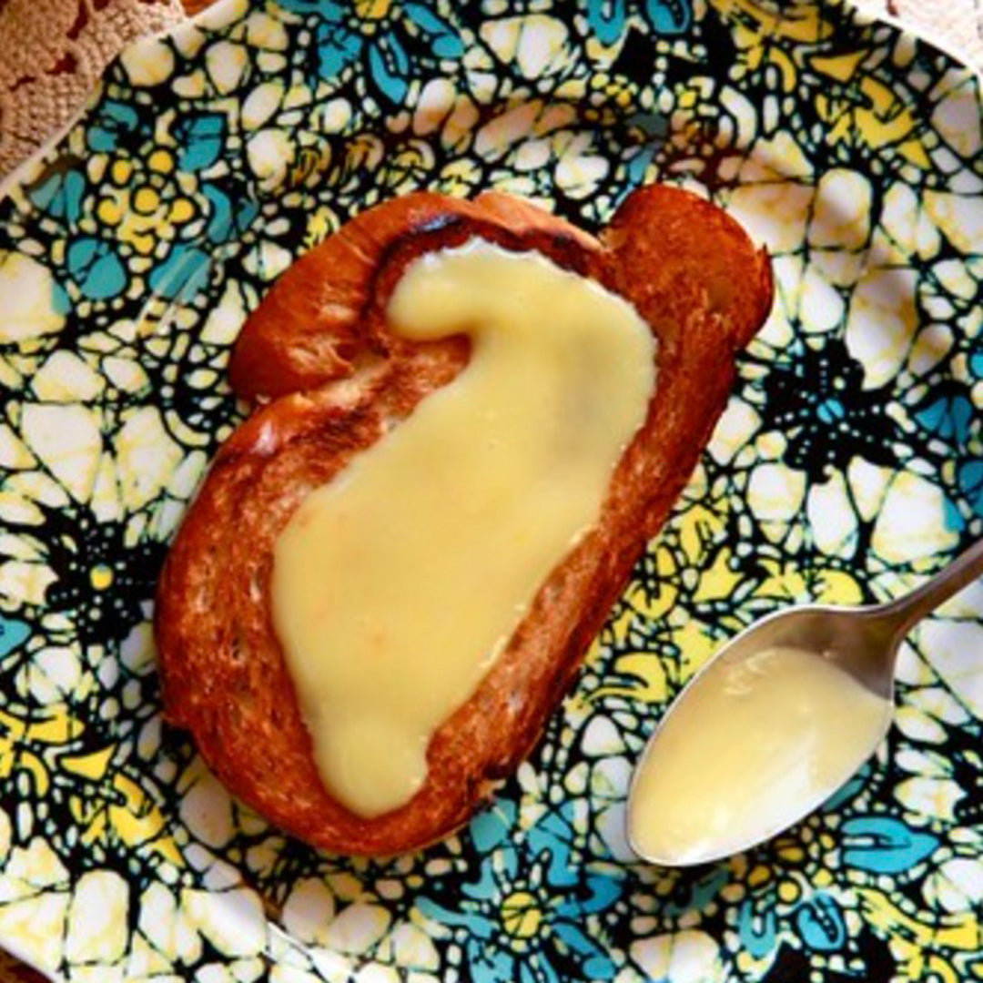 A slice of toasted bread topped with Tangerine Whipped Honey sits on a patterned plate beside a spoonful of the creamy yellow spread.