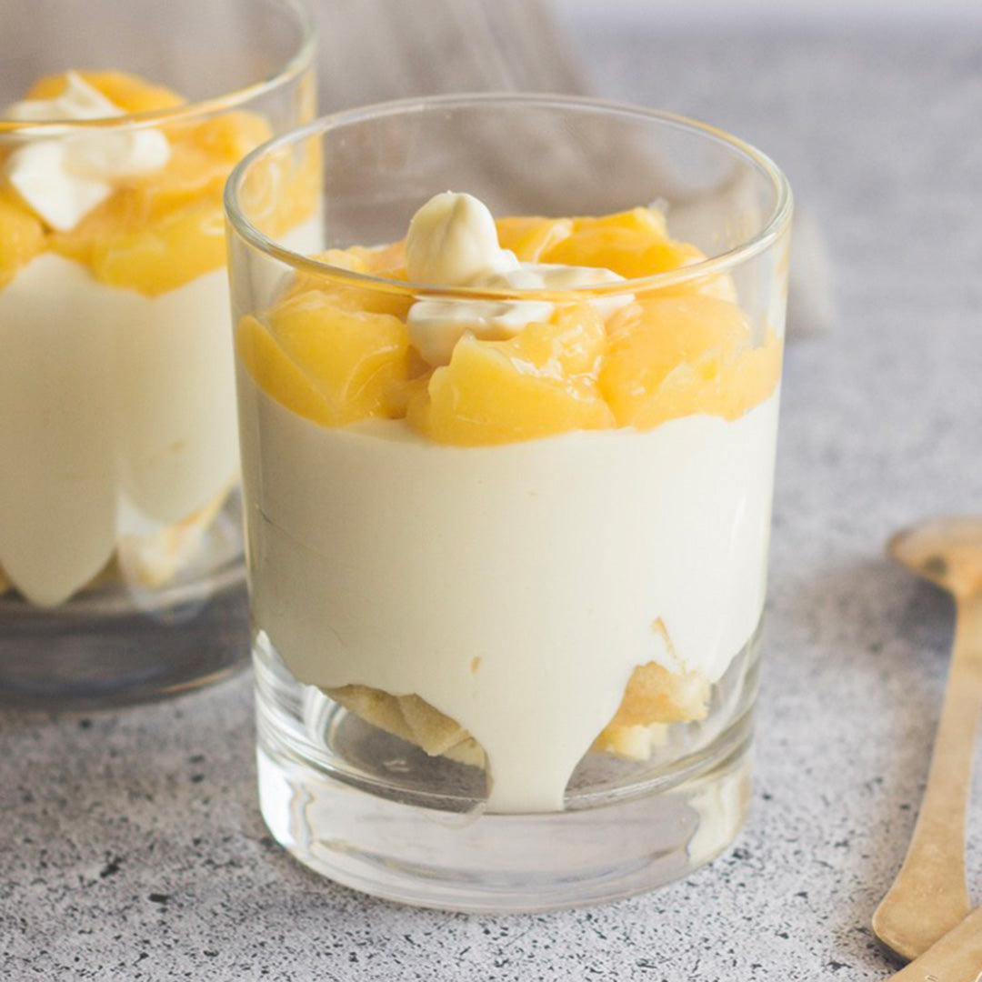 A glass filled with creamy white dessert, topped with yellow fruit chunks and a dollop of cream drizzled with Tangerine Whipped Honey, sits on a speckled surface. Another similar dessert is partially visible in the background.