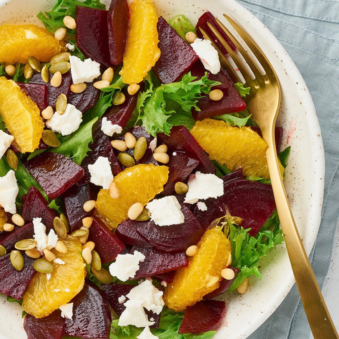 A salad of mixed greens, sliced beets, orange segments, crumbled feta, pumpkin seeds, and pine nuts, finished with a drizzle of Tangerine Whipped Honey and served on a white plate with a gold fork.