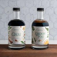Two bottles of Spiced Tea Exchange Chai and London Fog on a wooden surface with a tiled wall background.
