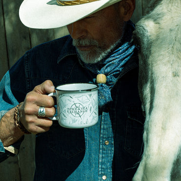 A man in a white cowboy hat, denim shirt, and blue bandana holds a white enamel mug printed with 'Spiced Tea Exchange' and leaf illustrations. A large white animal body is visible at the right edge, and a wooden fence is in the background.