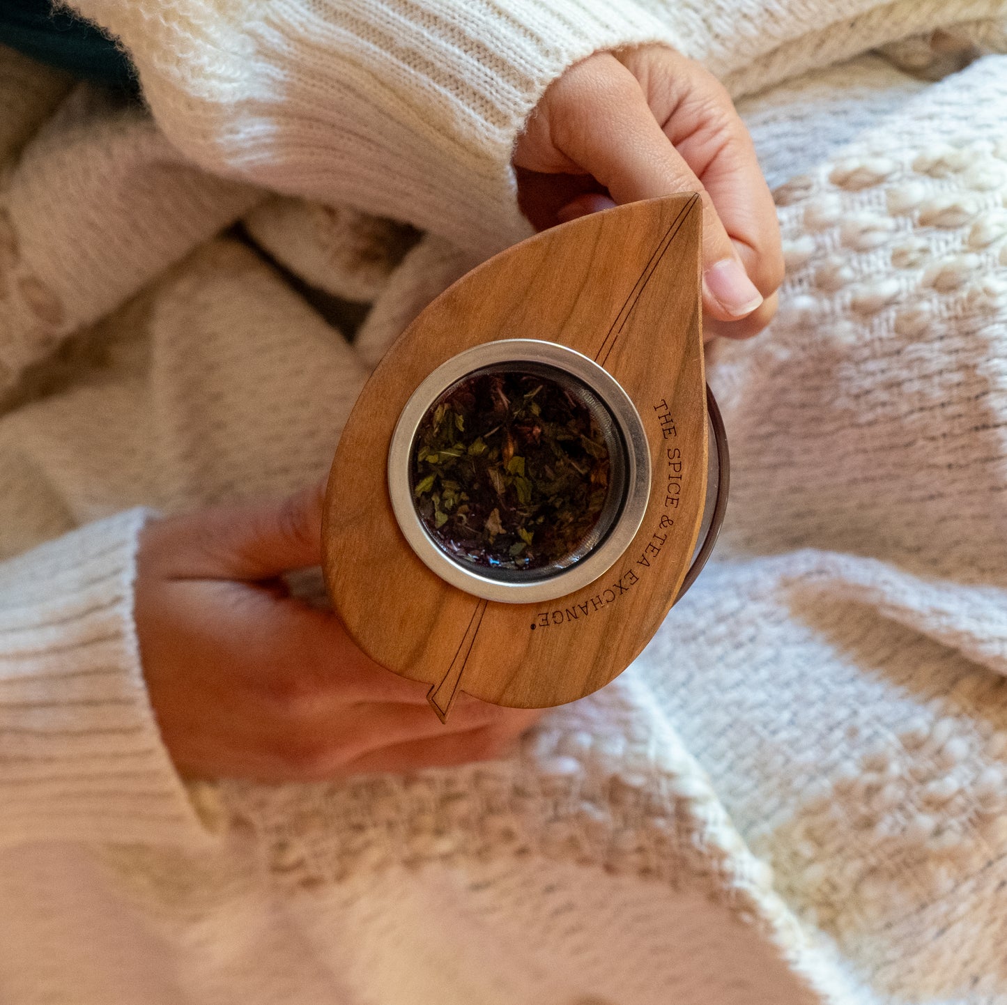 Wearing a cozy cream sweater, someone holds THE SPICE VALLEY EXPERIENCE wooden leaf-shaped container filled with loose tea leaves, next to a handcrafted Tea Nest Infuser (Vine, Leaf).