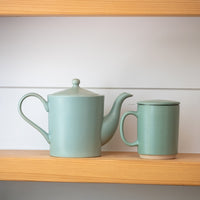 A light sage green ceramic teapot and matching mug with lids, displayed on a wooden shelf against a white paneled wall.