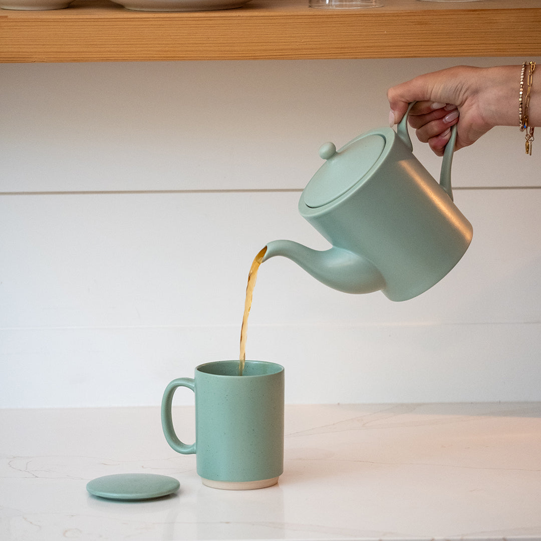 A sage green ceramic teapot with its lid off, being used to pour tea into a matching sage green ceramic mug on a white countertop.