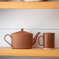 A matte terracotta brown ceramic teapot and matching mug with a lid, displayed on a light wood shelf against a white paneled background.