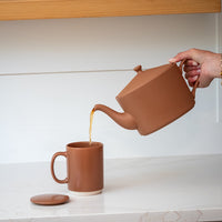 A matte terracotta brown ceramic teapot and pouring into a matching mug with a lid, displayed on a light wood shelf against a white paneled background.