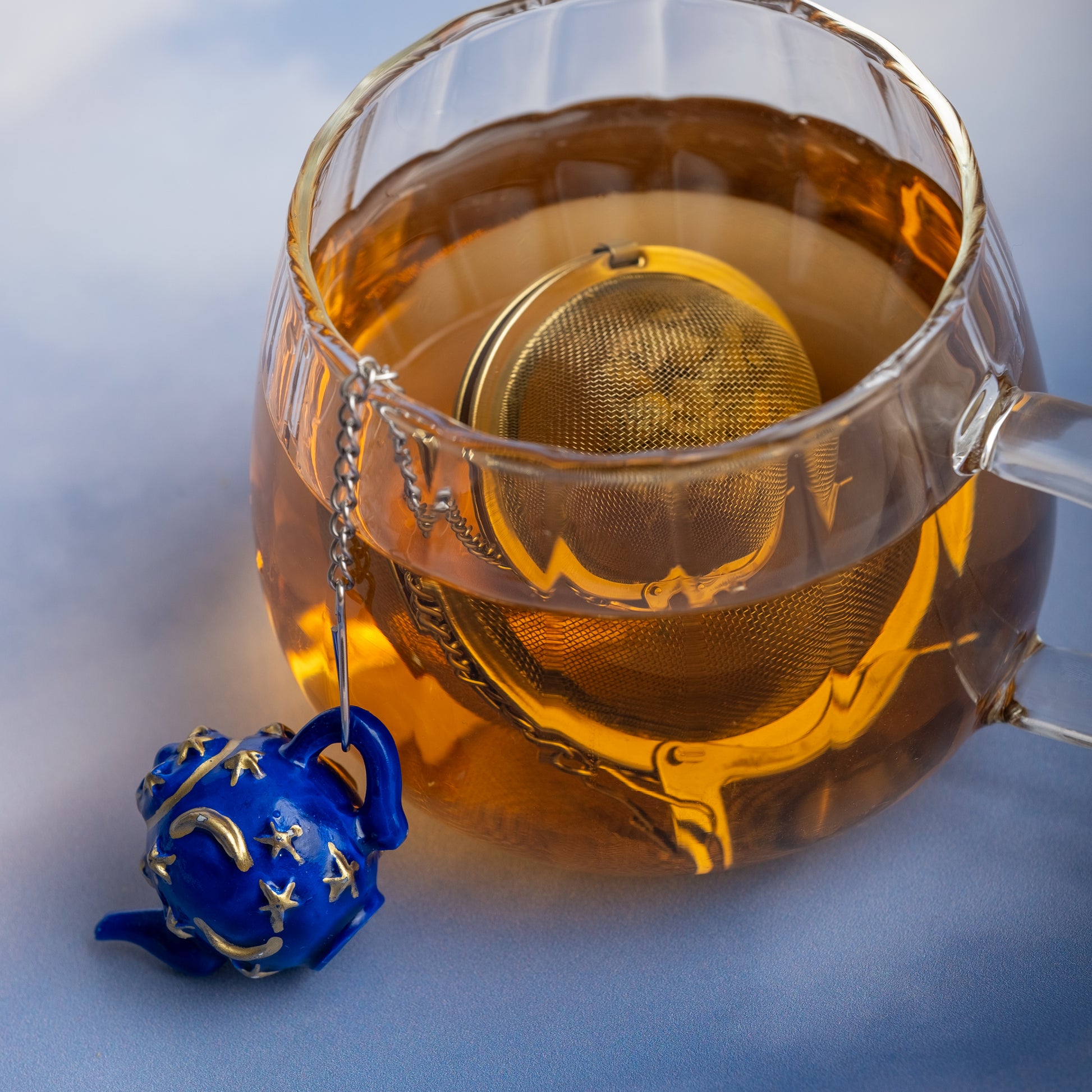 A glass cup of tea with the Infuser with Teapot Charm—a loose leaf infuser on a chain, adorned by a blue teapot-shaped charm featuring yellow stars—makes for a charming travel tea accessory beside the cup.
