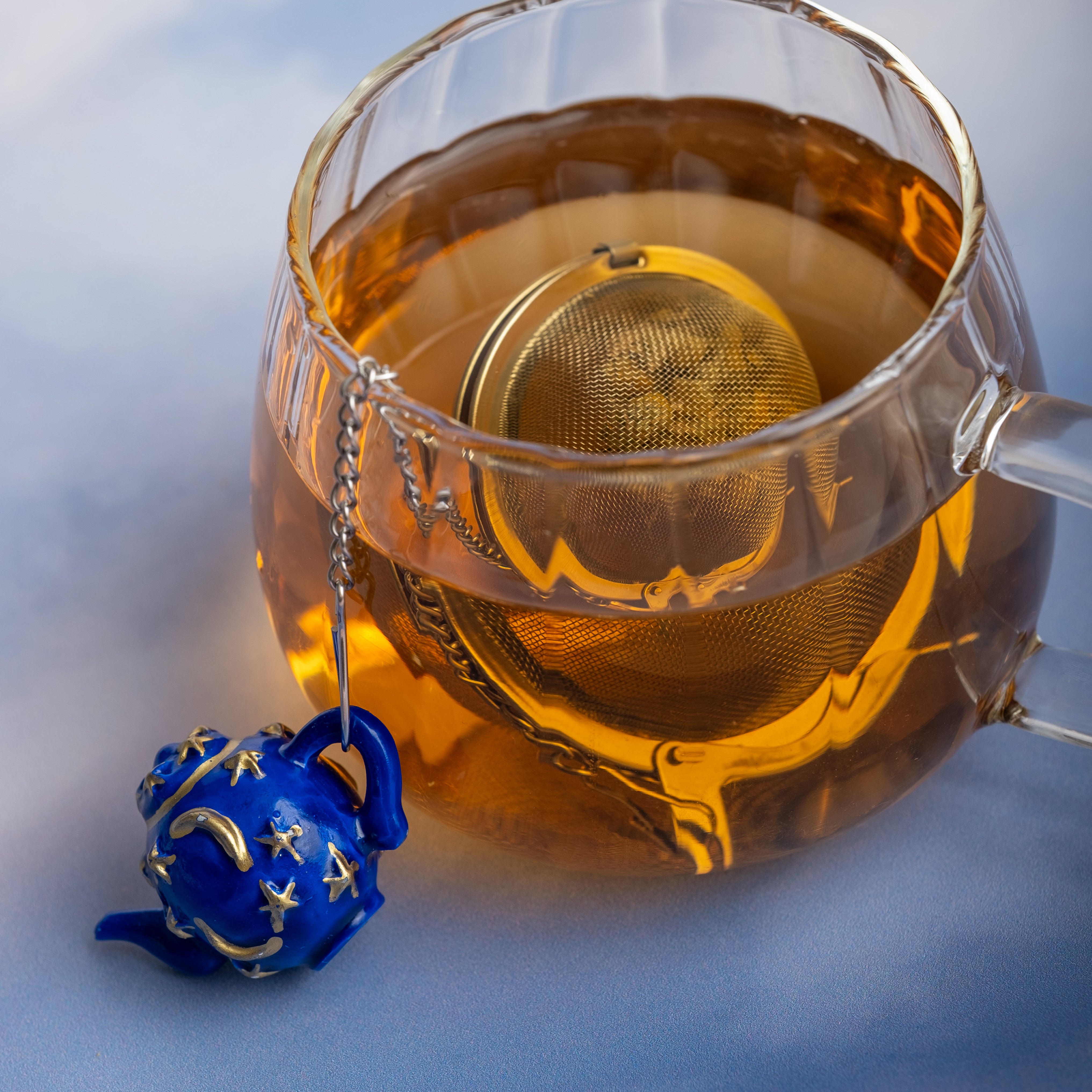 A glass cup of tea with the Infuser with Teapot Charm—a loose leaf infuser on a chain, adorned by a blue teapot-shaped charm featuring yellow stars—makes for a charming travel tea accessory beside the cup.