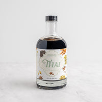 A bottle of Spice Tea Exchange Thai Leaf Concentrate, featuring a clear glass bottle with dark liquid inside and a white label decorated with tea and spice illustrations, set on a light marble surface against a white background.