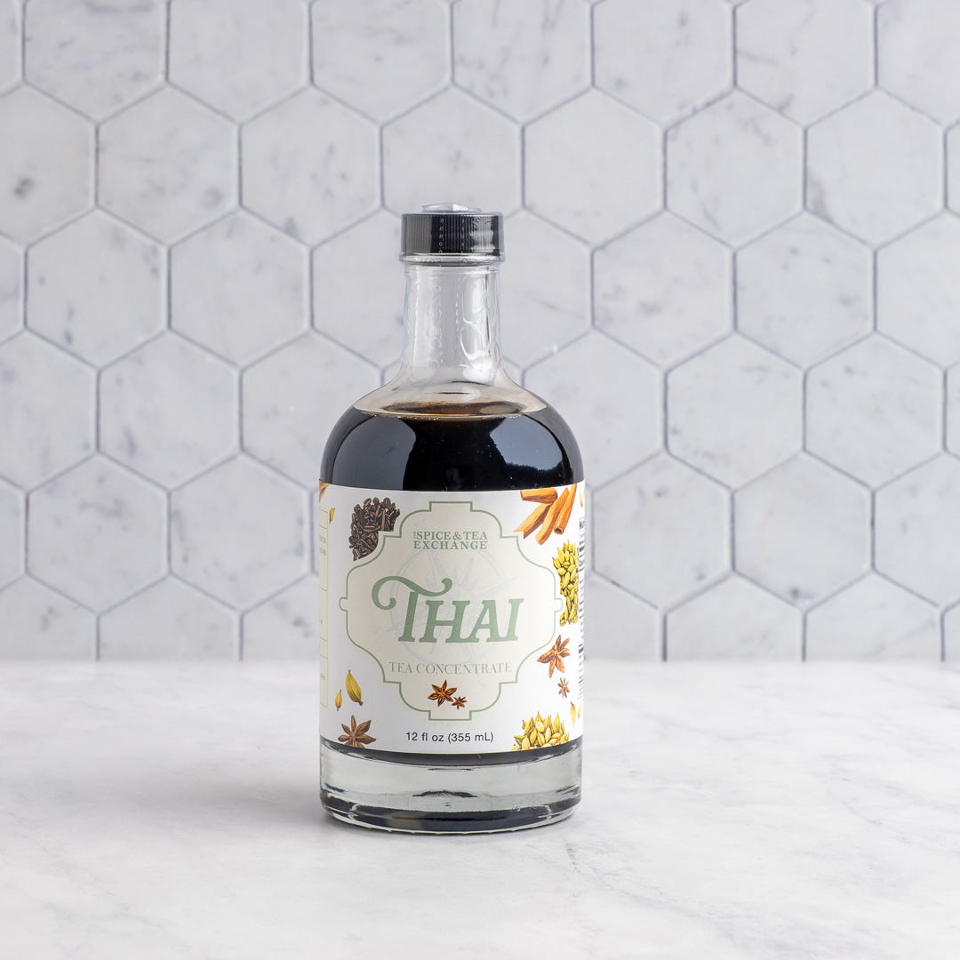 A bottle of Thai Tea Concentrate on a white marble surface against a white hexagonal tile wall. The bottle is clear glass, showing the dark coffee inside, and has a cream-colored label with green and gold lettering and illustrations of spices.