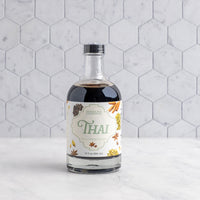 A bottle of Thai Tea Concentrate on a white marble surface against a white hexagonal tile wall. The bottle is clear glass, showing the dark coffee inside, and has a cream-colored label with green and gold lettering and illustrations of spices.