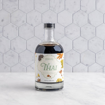 A bottle of Thai Tea Concentrate on a white marble surface against a white hexagonal tile wall. The bottle is clear glass, showing the dark coffee inside, and has a cream-colored label with green and gold lettering and illustrations of spices.