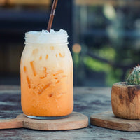 An iced thai tea drink in a clear glass mason jar with a metal straw, sitting on a wooden coaster on a rustic table. A small potted cactus is visible on the right, with a blurred outdoor background.