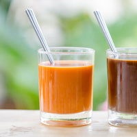 Two clear glass cups with metal straws filled with thai iced tea (one with cream), set on a light surface with a soft blurred green background.