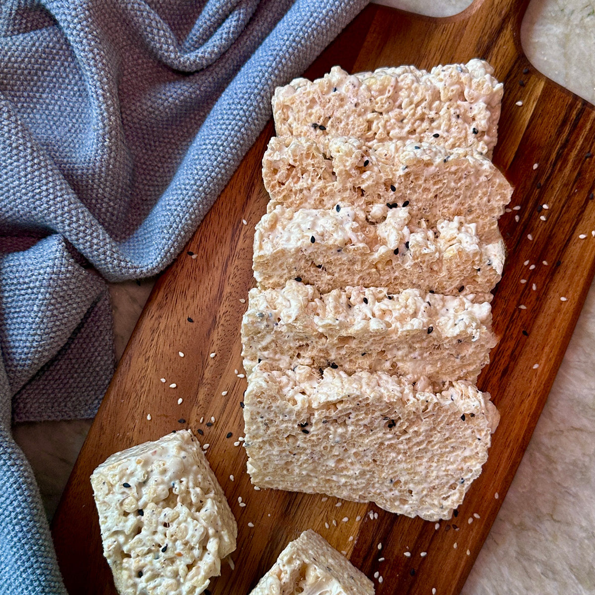 Four rectangular slices of crispy rice treat with visible black sesame seeds, arranged on a wooden board with a blue-gray cloth in the background.