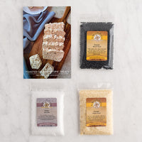 Three packaged baggies or product arranged on a light marble surface with a printed card showing toasted sesame crisp treats.