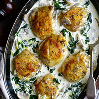 Overhead close-up of baked chicken thighs in a creamy mushroom and spinach sauce in an oval baking dish, with a metal serving spoon on the right.