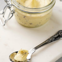 A close-up of a creamy pale yellow  butter in a small clear glass jar with a metal clamp lid, with a spoonful of the product in the foreground on a light stone surface.