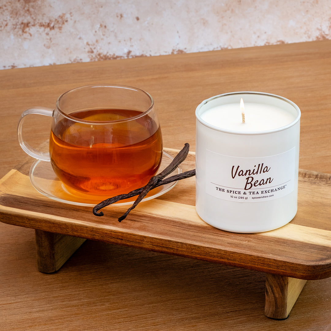 A white candle labeled 'Vanilla Bean' sits on a wooden stand beside a glass cup of tea and vanilla beans on a wooden table. .