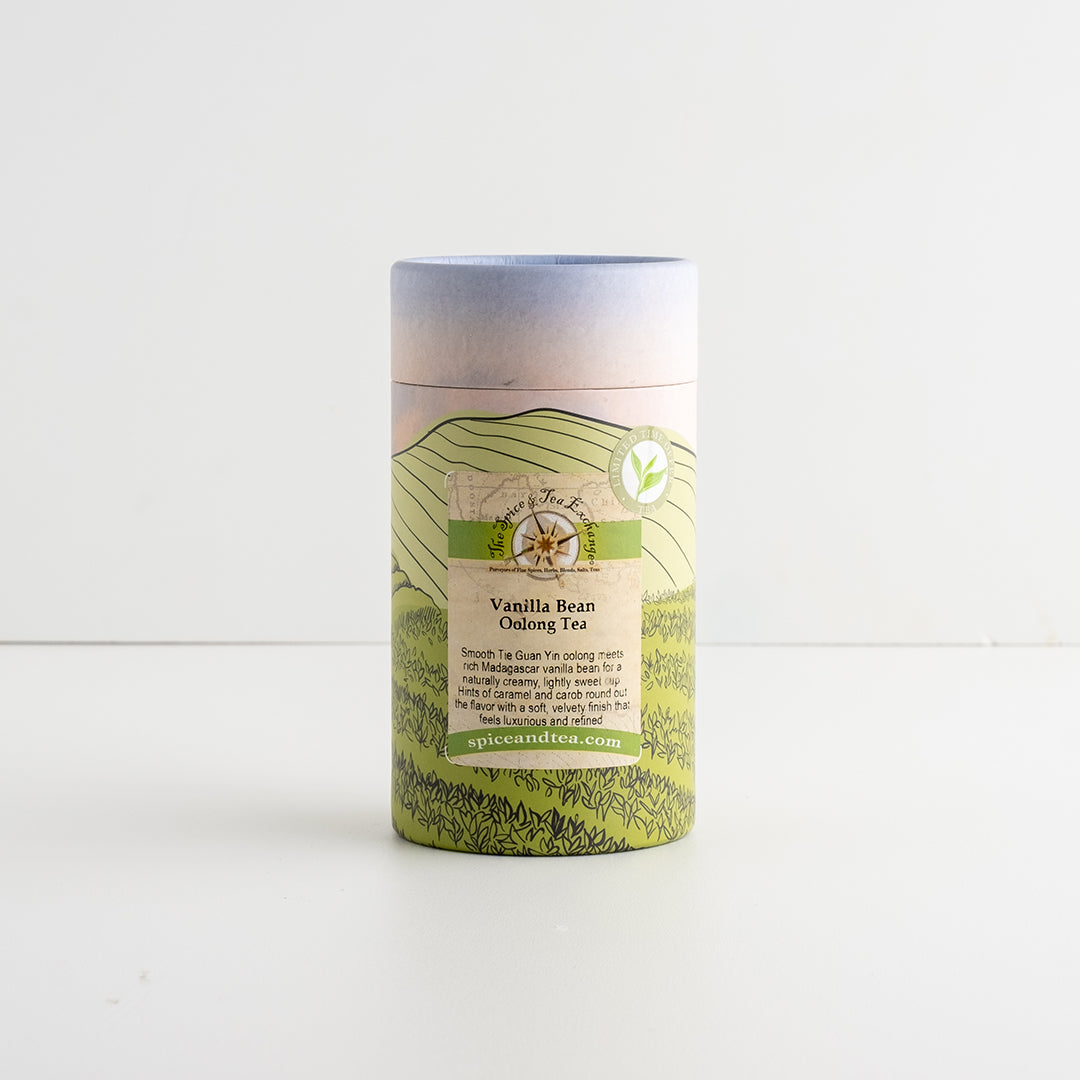A cylindrical paperboard canister of Vanilla Bean Oolong Tea, featuring a green and beige landscape illustration and a light blue lid, set against a plain white background.