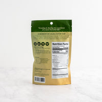 The back of a stand-up pouch package for Vanilla Matcha Latte, featuring a green top section with a quote and product icons, and a beige lower section displaying the Nutrition Facts label, photographed on a white marble surface against a white background.