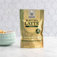 A stand-up pouch of Spice Tea Exchange Matcha Latte Vanilla tea mix on a white marble surface, with a small pale green bowl and wooden spoon partially visible at the left edge against a white hexagonal tile backsplash.