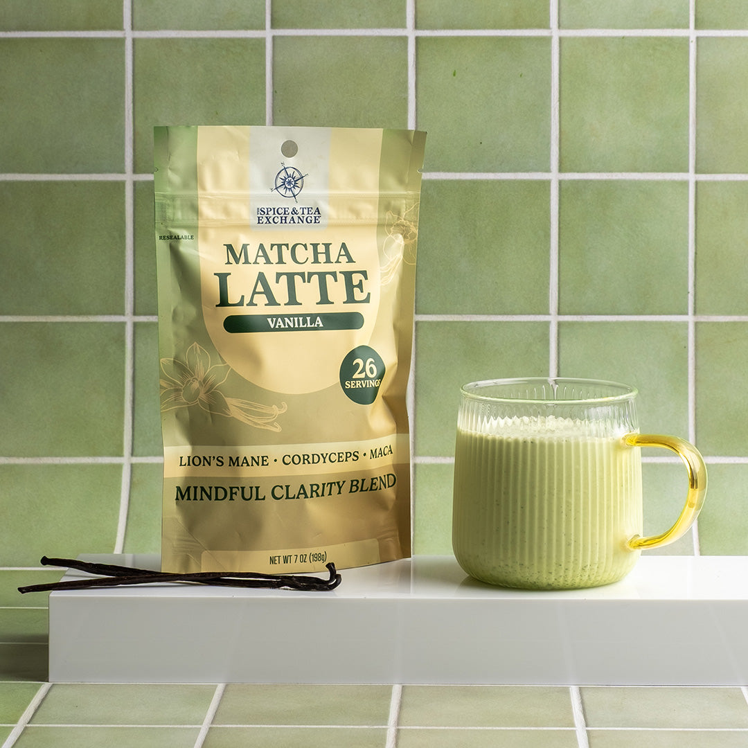 A stand-up pouch of Matcha Latte Vanilla herbal tea by Spice & Tea Exchange sits on a white shelf beside a ribbed glass mug filled with pale green tea, with vanilla beans in front, against a green tiled wall.