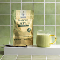 A stand-up pouch of Matcha Latte Vanilla herbal tea by Spice & Tea Exchange sits on a white shelf beside a ribbed glass mug filled with pale green tea, with vanilla beans in front, against a green tiled wall.