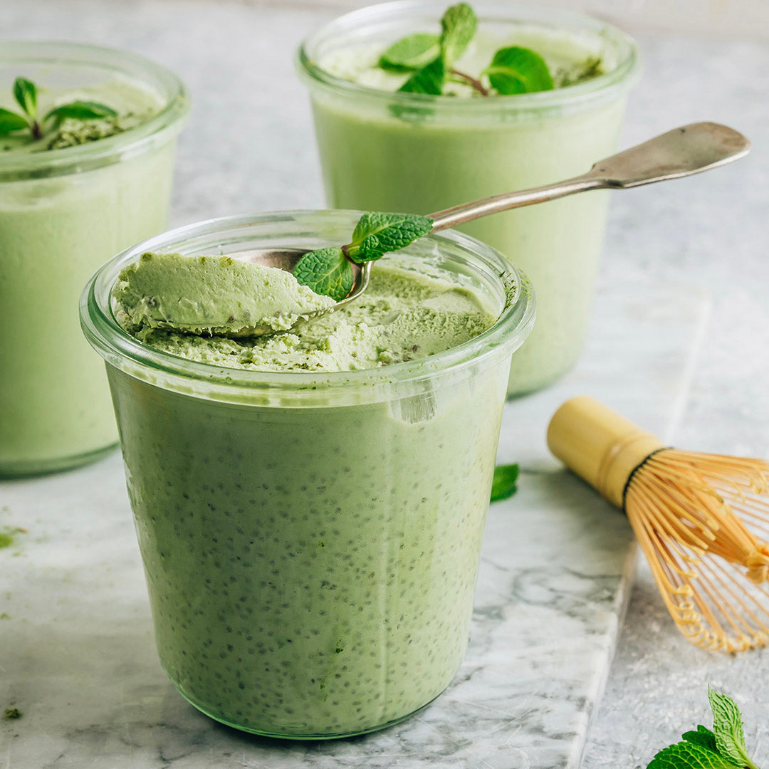 Three clear glass jars filled with light Vanilla Matcha Latte chia pudding, arranged on a marble surface with a metal spoon resting in the front jar and a bamboo whisk nearby.
