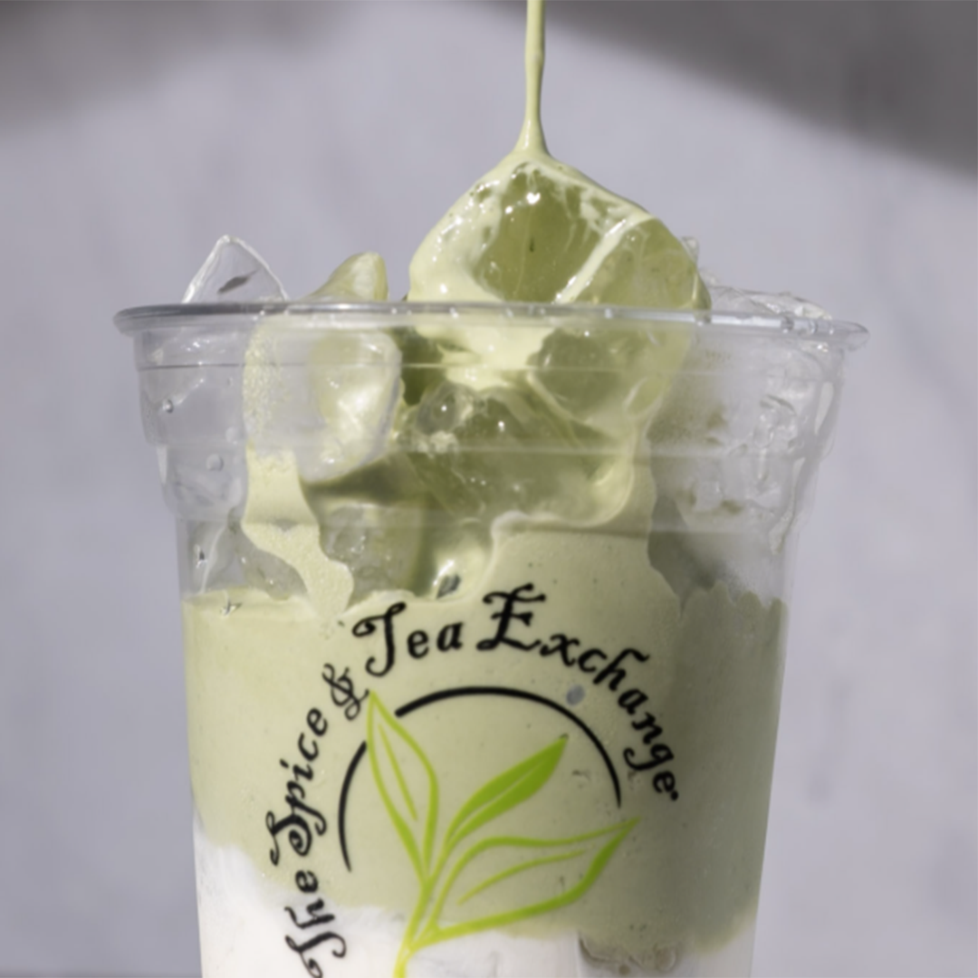 A clear plastic cup filled with a light green iced Vanilla Matcha Latte and ice cubes, being poured. The cup has a printed logo reading 'The Spice & Tea Exchange' with a green leaf graphic.