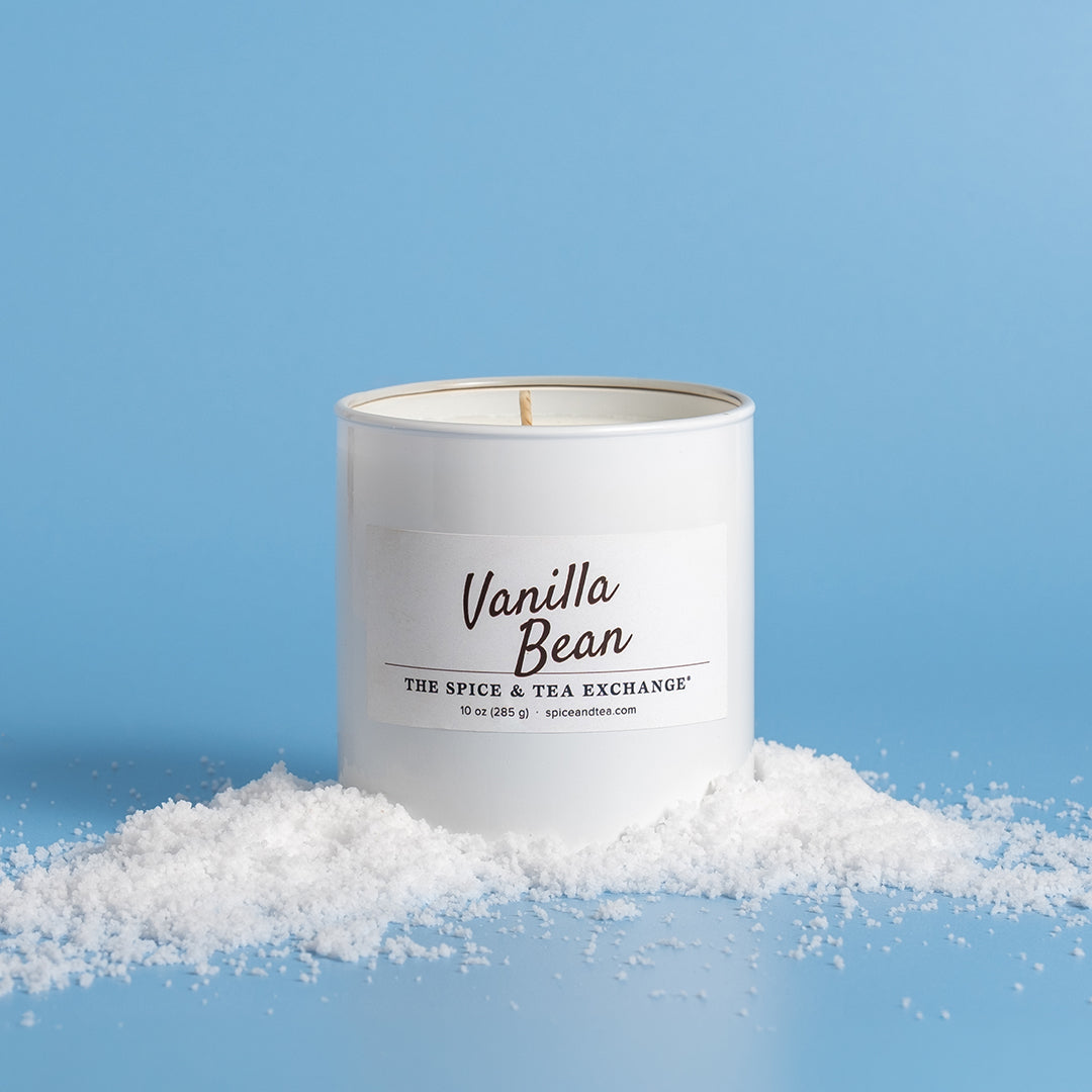 A white glass candle jar labeled 'Vanilla Bean' by The Spice & Tea Exchange, styled with white snow on a light blue background.