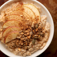 A top-down close-up of a white bowl filled with oatmeal topped with sliced apple, granola, and a dusting of cinnamon.