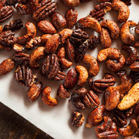 A close-up overhead view of assorted roasted nuts, including cashews, pecans, almonds, and walnuts, coated in a glossy brown sugar and spice mixture on parchment paper.