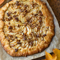 A round apple tart with a rustic golden-brown crumb crust, filled with sliced apples and topped with chopped walnuts, served on parchment paper.