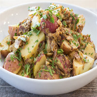 A white bowl filled with chunky red potato salad, garnished with chopped parsley and mixed with a savory, brown seasoning. The dish sits on a wooden surface.
