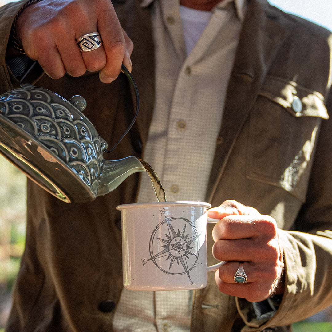 A person in a brown jacket pours hot water from a patterned metal kettle into a white enamel mug with a compass design, shown in a warm outdoor setting.