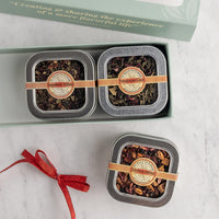 Two square metal tins of loose-leaf herbal tea displayed on a light marble surface, with one tin inside an open pale green gift box and the other placed below. Each tin has a tan paper band and a round label.