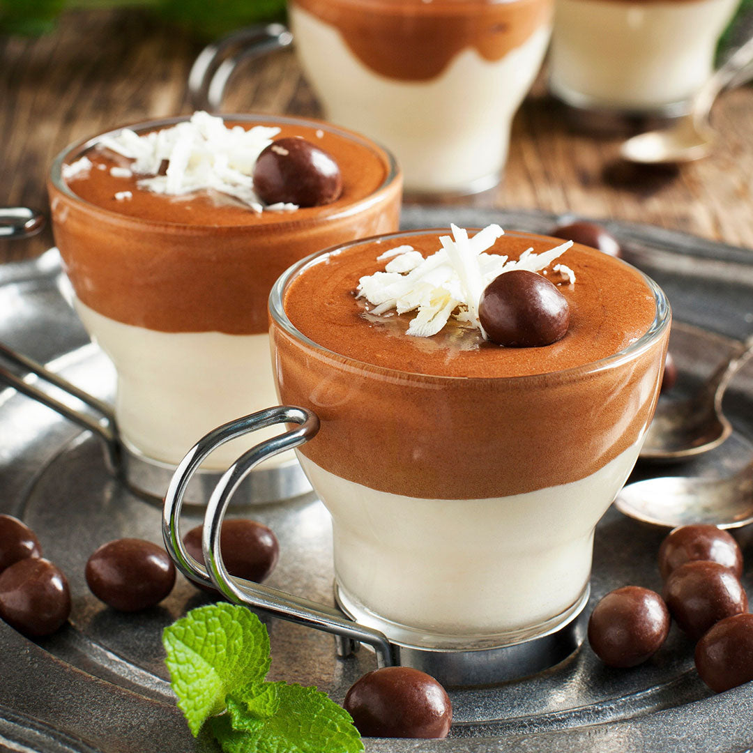 Two glass cups filled with layered dessert on a metal tray with chocolate morsels and mint leaves.