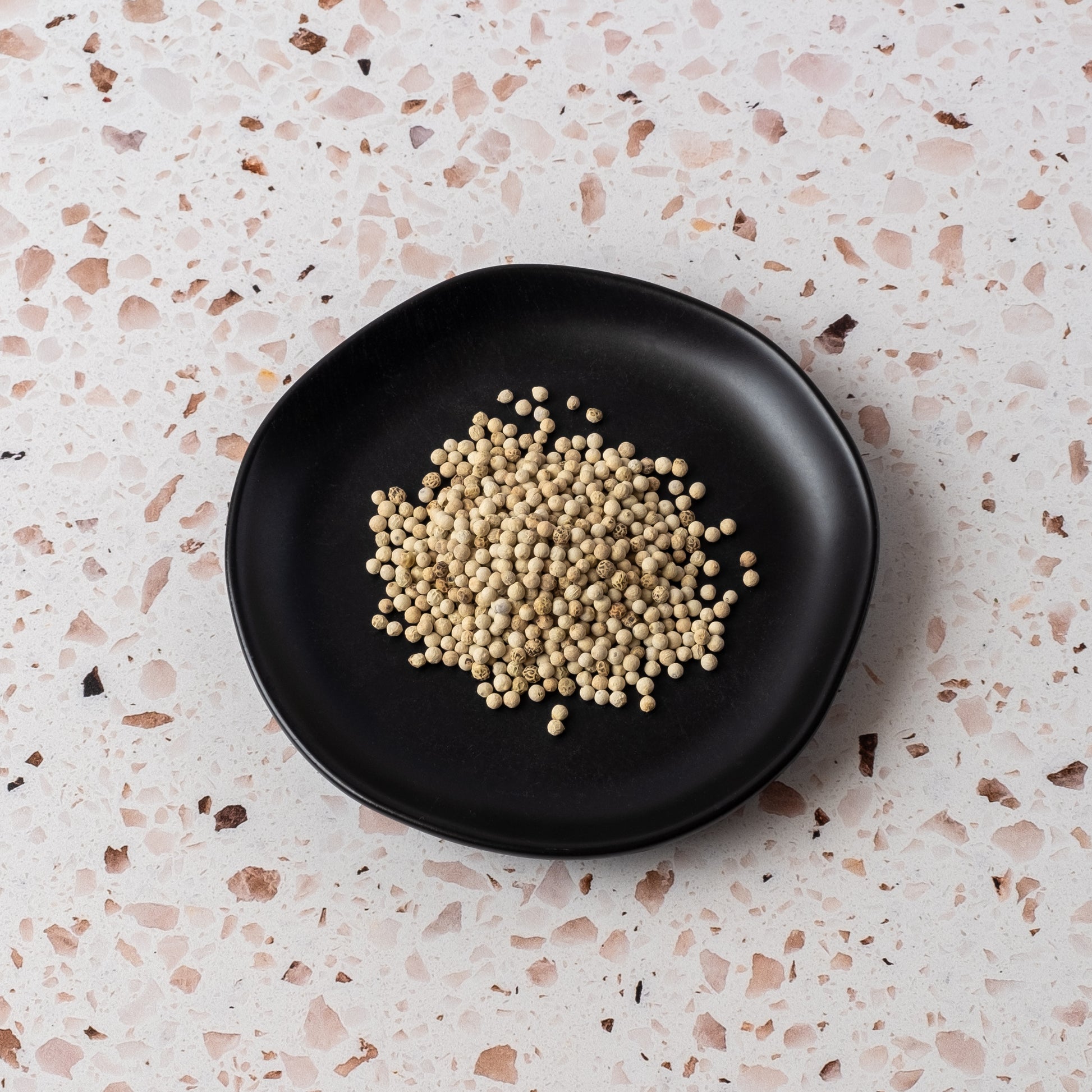 A small pile of White Peppercorn, a prized spice for cream sauces, sits on a round black plate placed on a light speckled terrazzo surface.