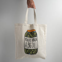A hand holds The Spice & Tea Exchange "You're Kinda A Big Dill Tote Bag," which features smiling pickles in a jar with the text “You’re Kinda a Big Dill” on a plain light gray background.